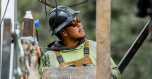Construction worker wearing a hard hat and safety glasses looks up at equipment on a construction site, holding a metal mesh panel.